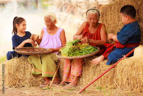Fotografia Traditional Asian Thai rural daily life, grandchildren in cultural costumes help their seniors preparing local food ingredients for the meal