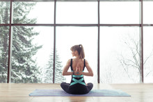 Back View Of Healthy Young Woman Practicing Yoga And Sitting In Padmasana Yoga Pose At Training Hall Or Home Near A Windows