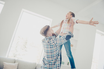 Sticker - Low angle view of strong stylish father raising up his glad excited laughing daughter holding hands to the side enjoying game spending time in house white living room