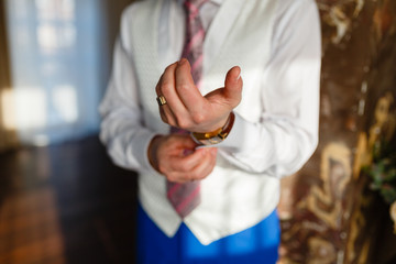 Wall Mural - The man fixes the cuffs on the white shirt and stylish vest and blue pants with expensive gold accessories, watches, rings and rings. The businessman is preparing to meet in an expensive interior.