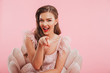 Portrait closeup of flirty winking woman in dress sitting in armchair and pointing finger at you, isolated over pink background