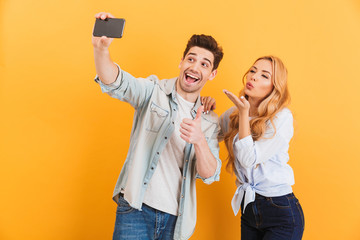 Wall Mural - Portrait of two attractive people man and woman taking selfie photo on cell phone while gesturing at camera, isolated over yellow background