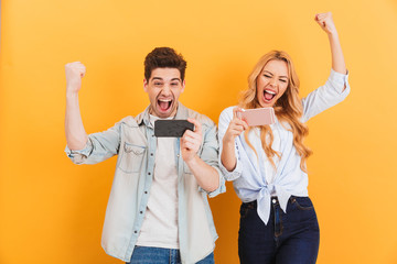 Wall Mural - Image of ecstatic man and woman rejoicing while playing together video games on mobile phones, isolated over yellow background