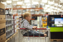Bored Young Boy Stares As He Sits In The Top Basket Of A Metal Shopping Trolley In A Large Store.
