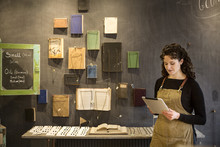 Woman With Curly Brown Hair Wearing Apron Standing In Her Pottery Shop, Looking At Digital Tablet, Display Of Books And Jewellery.