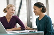 © Erickson Stock - Two smiling women sitting next to each other at a table inside an office.