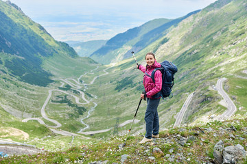 Wall Mural - Happy female tourist pointing with walking stick to beautiful view of magnificent winding Transfagarashan road in Romania. Woman climber green hills mountains happy rock travel route path