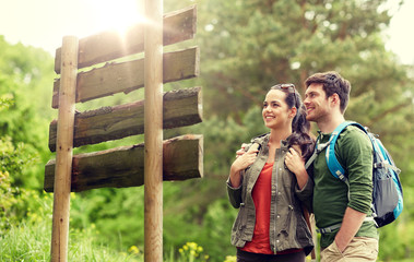 Poster - adventure, travel, tourism, hike and people concept - smiling couple with backpacks looking at signpost outdoors