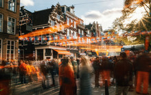 Streets Of Amsterdam Full Of People In Orange During The Celebration Of Kings Day. Blurred People At Sunset With Sunlight And Orange Decorations.