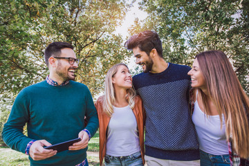 Wall Mural - Image of four happy smiling young friends walking outdoors in the park holding digital tablet