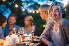 Group Of Friends Gathered Around A Table In A Garden On A Summer Evening To Share A Meal And Have A Good Time Together. Focus On A Beautiful Woman
