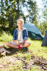 Wall Mural - Little backpacker with cup of tea sitting on grass by campfire on background of his tent