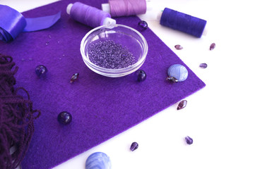 A set of materials for needlework in violet color on a white background. Beads, threads, a satin ribbon and wool for knitting.