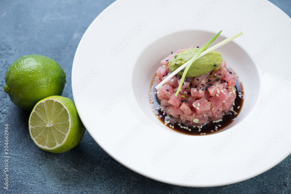 White plate with tuna tartar in soy sauce topped with guacamole quenelle and green onion, studio shot