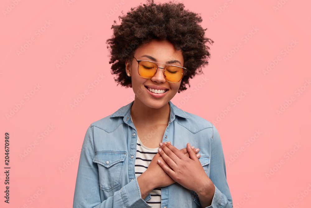 Friendly looking female with Afro hairstyle, keeps hands on chest ...