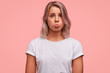 Abused pretty female curves lower lip, being offended during quarrel, looks with sorrowful miserable expression, wears casual white t shirt, isolated over pink background. Disappointed unhappy woman