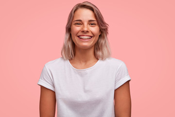 Sticker - Joyful teenage girl with dental braces on teeth, rejoices positive news, wears casual t shirt, isolated on pink background. Happy young hipster female with broad shining smile, rests indoor.