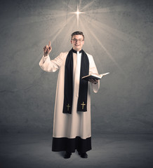 A young male priest in black and white giving his blessing in front of grey wall with glowing cross concept.