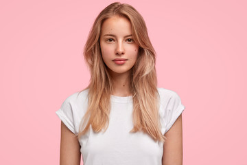 Wall Mural - Horizontal portrait of serious beautiful female with long hair, has no make up, looks delightfully at camera, wears casual white t shirt, poses against pink background. People, lifestyle, beauty
