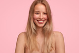 People, natural beauty and positive emotions concept. Pleased young female with long hair, poses nude against pink background, smiles joyfully as being photographed for popular fashion magazine