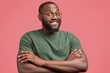 Wall Mural - Photo of attractive dark skinned male with glad expression, keeps hands crossed, rejoices achieving success in particular sphere, wears casual t shirt, round spectacles, isolated on pink background