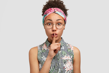 Wall Mural - Secret young African American housewife shows silence sign, asks to be silent as her child is sleeping, wears headband and blouse, isolated on white background. People and conspiracy concept
