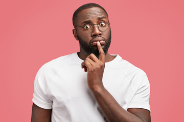 Wall Mural - Portrait of surprised stunned African American male worker, puzzled to realize his fault, stares through spectacles, has dark bristle, healthy pure skin, wears casual t shirt, model in studio