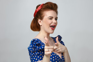 Close up shot of glamorous attractive young brunette female in vintage dotted dress blinking and pointing index fingers at camera, having mysterious look, posing against gray studio wall background