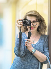 Adult happy smiling caucasian woman photographer standing next to a wall and taking a photo with photo camera outside at sunny day. Hobby or Photographer job