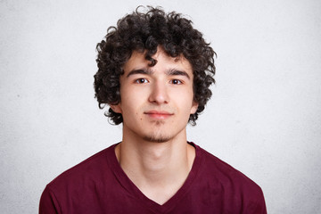 Close up shot of curly European male with little beard, wears casual sweater, isolated over white concrete wall, listens attentively interlocutor. Handsome teenager boy with crisp dark hair.