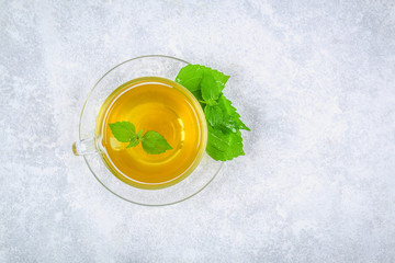 Leaves of fresh green nettle and a clear glass cup of herbal nettle tea on a gray concrete table. Top view.