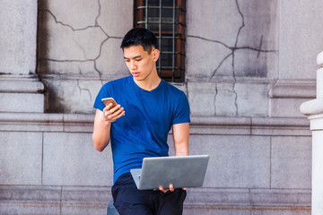 Young Asian American Man studying, working in New York, wearing blue T shirt, black pants, sitting on pillar on old street on campus, working on laptop computer, looking down, texting on cell phone..