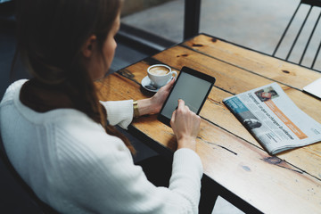 View from back photo of young caucasian female surfing the web on a digital tablet while sitting with a morning coffee cup and newspaper at the wooden table in a coffee shop. Woman reading news online