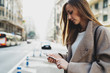Portrait of beautiful designer girl looking at mobile phone while chatting online on a blurred urban background. Stylish hipster girl reading messages on smartphone while crossing the road