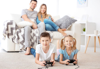 Wall Mural - Cute children playing video games while parents resting on background