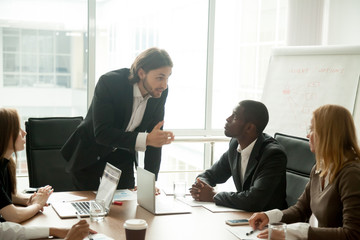 Wall Mural - Angry mean boss scolding incompetent employee for bad work results at diverse group meeting, company executive reprimanding admonishing office worker for business mistake at multiracial team briefing