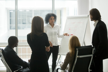 Serious african female leader or coach gives presentation of new marketing plan at sales team meeting, black employee explains colleagues new client management strategy idea at group office training