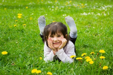 Happy little girl lying down on the grass with hands holding her face and feet up at park full of yellow flowers. Cute kid smiling on spring season