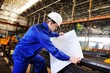 © Evgeniy Kalinovskiy - Working engineer in a white construction helmet with a project or drawing plan on the background of an industrial plant
