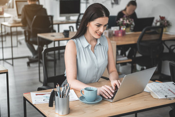 Poster - Portrait of happy young businesswoman typing in laptop while locating at desk. Cheerful worker concept