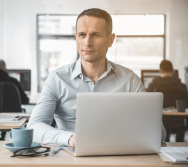 Poster - Portrait of pensive worker typing in notebook computer while sitting at table opposite colleagues in office. Contemplative employer during labor concept