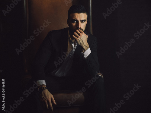 Fotomural A handsome man with a beard and in a classic suit sits in a leather chair and looks at the camera