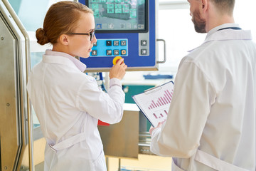 Wall Mural - Beautiful technician and her bearded colleague wearing white coats adjusting equipment while standing at production department of modern plant