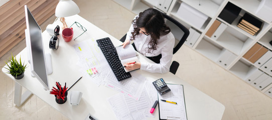 A young girl is sitting at the computer desk in the office, holding a pen in her hand and looking at the notepad.