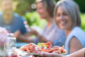 Close-up on a hand serving a dish of dry ham and cheese to friends gathered around a table in a garden to have fun together.