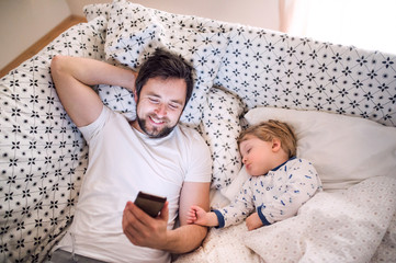 Wall Mural - Father with smartphone and a sleeping toddler boy in bed at home.