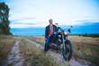 © lkoimages - Young biker wears black Leather Jacket and jeans, sits on his black old motorbike, holds white helmet and looks on beautiful soft sunset
