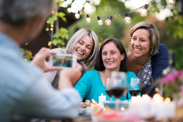 One summer evening friends gathered around a table in the garden for a good time. A man takes a picture of three female friends in their forties