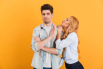 Portrait of young displeased man gesturing to stop with hand while beautiful woman kissing his cheek, isolated over yellow background