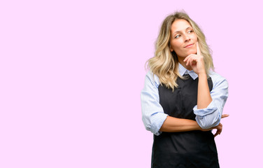 Young shop owner wearing black apron thinking and looking up expressing doubt and wonder